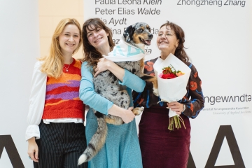 Drei Personen, eine mit Hund auf dem Arm, vor dem Transparent mit der Namensliste
                                          aller Absolvent*innen