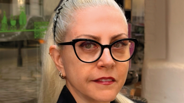 A portrait photograph of Basak Senova with
                                          blonde hair tied back and wearing black glasses. She is wearing a black jacket and a necklace, and is looking directly into
                                          the camera. In the background, an exhibition space with glass panels and works of art can be seen.