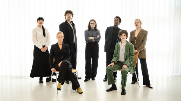 A group of 7 people is standing in front of a white background and is posing for the camera