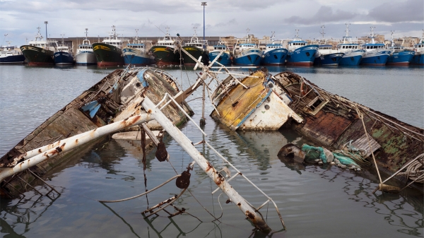 Plakat zur Ausstellung „Soil & Water:
                                          Mediterranean Crossing“ mit einem Foto von versunkenen, beschädigten Fischerbooten im Vordergrund und einer Reihe intakter
                                          Boote im Hafen im Hintergrund. Das Foto stammt aus der Serie Hotspot Mediterraneo (2025) von Francesco Bellina. Courtesy of
                                          the artist.