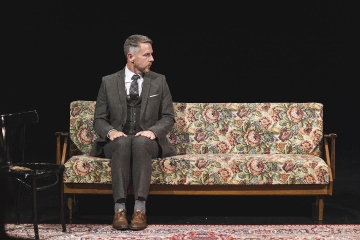 Der Performer Jim Libby auf einem Sofa auf einer dunklen Bühne